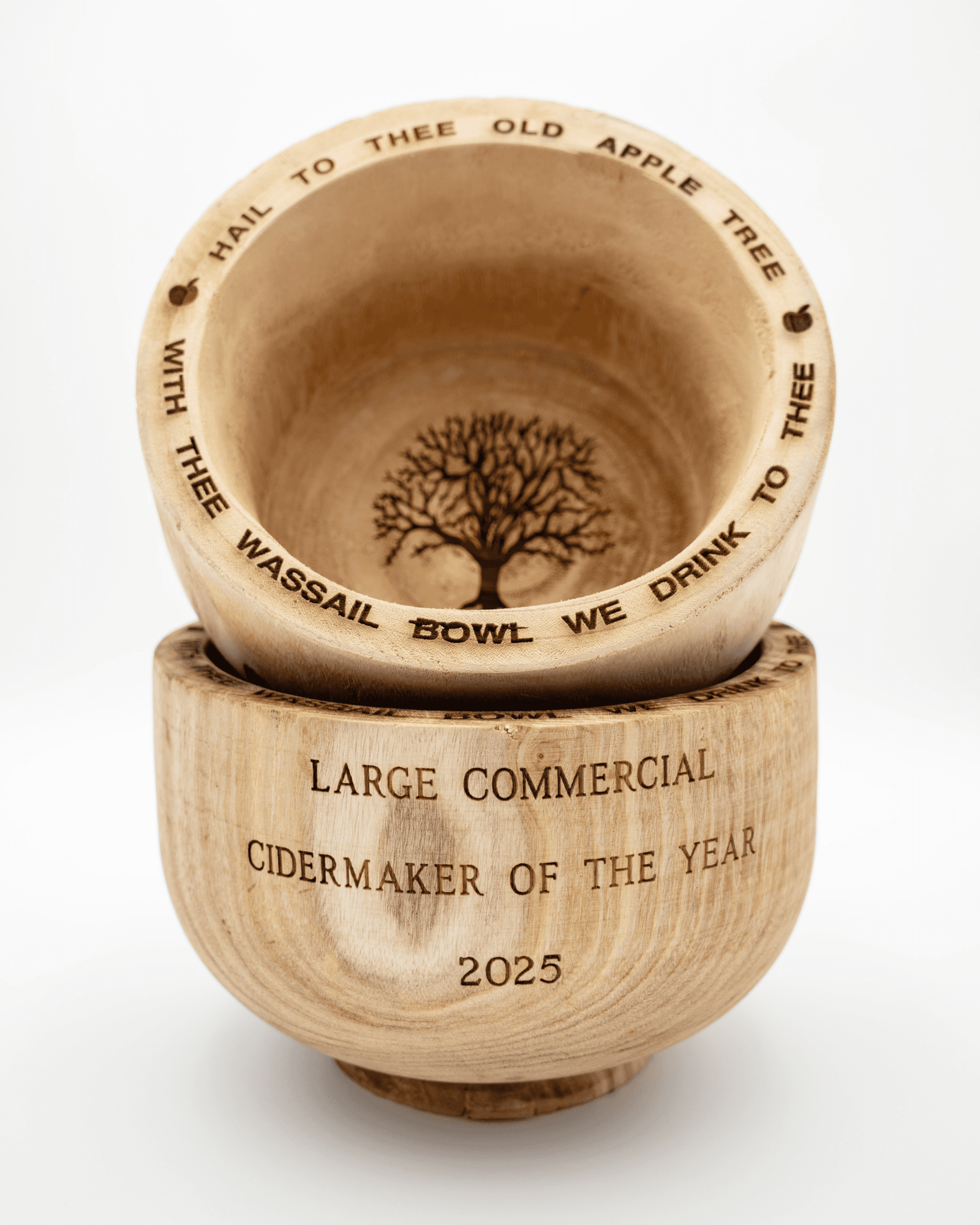 Two wooden bowls, with one sitting inside of the other. The bottom bowl reads "large commercial cider maker of the year 2025." The second bowl reads "Hail to thee old apple tree with thee wassail bowl we drink to thee." The top bowl also has an image of a tree carved into it.