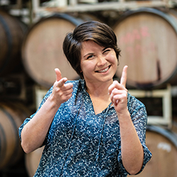 Madison smiles at the camera and shoots finger guns with a background of barrels behind her. Celebrating International Women's Day