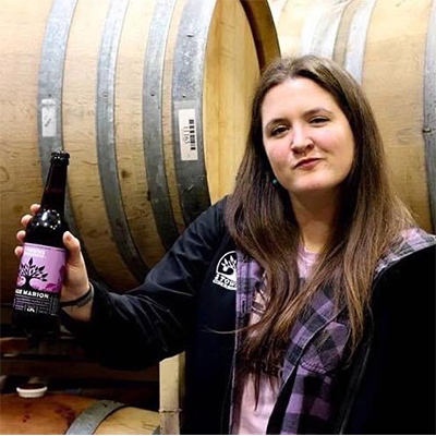 Amanda smiles while holding a Made Marion in her hand in front of barrels at the ciderhouse. Celebrating International Women's Day