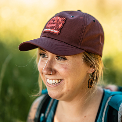 Megan smiles off to the left of the camera, wearing a Prickly Pearadise maroon hat with a blurry green background behind her. Celebrating International Women's Day